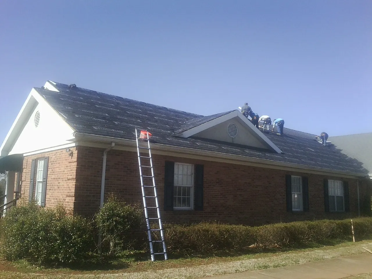 Crew tearing shingles off a brick ranch roof with a ladder set — Emergency Roof Repair work by our crew in Pointblank