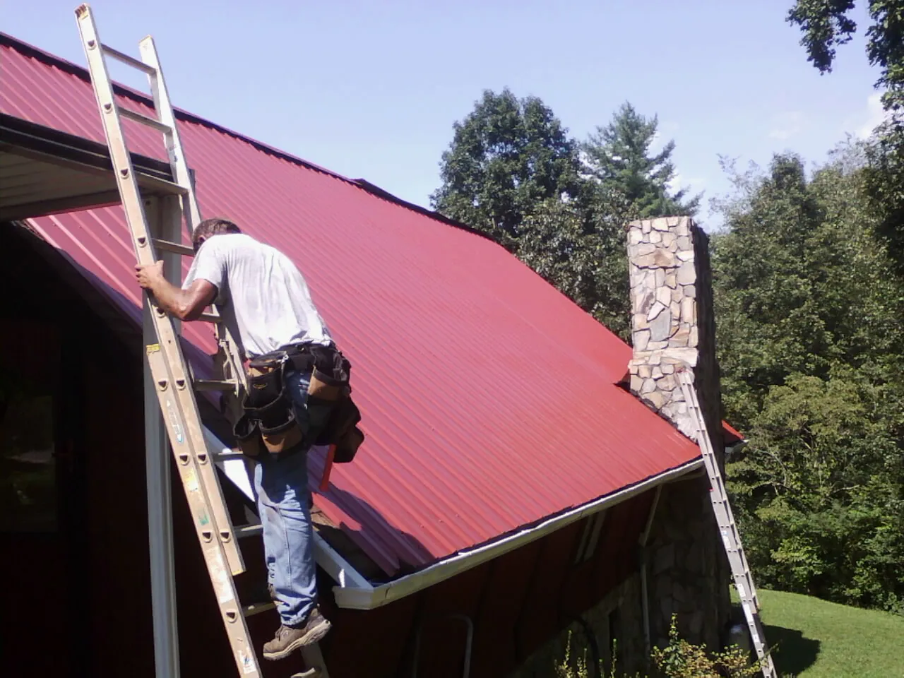 Roofer on a ladder beside a red metal roof and stone chimney — Soffit & Fascia Repair by Roofing Brothers in Pointblank